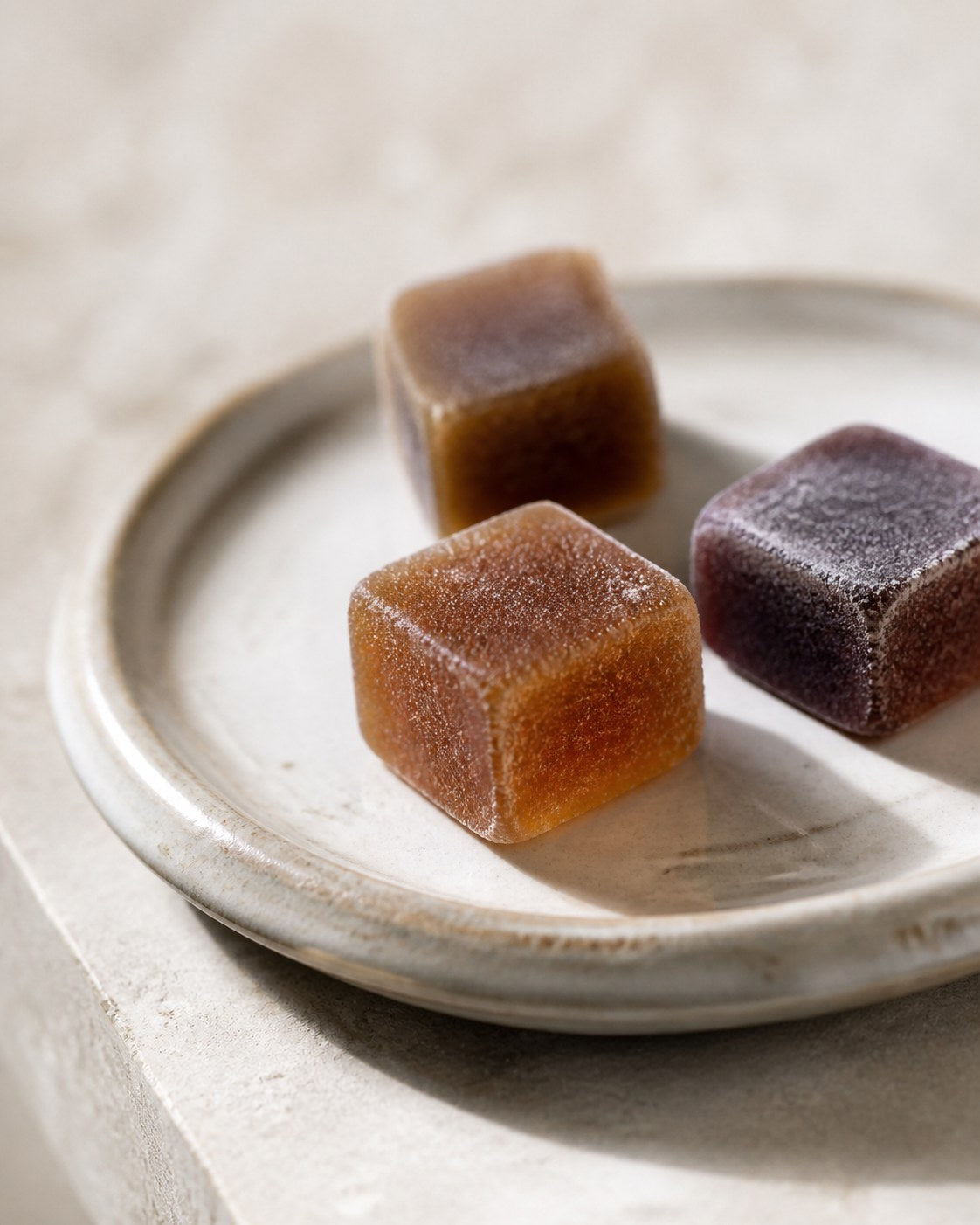 Three Veya cube gummies on a small stoneware dish, colour varying from amber to deep plum, matte dusted surface